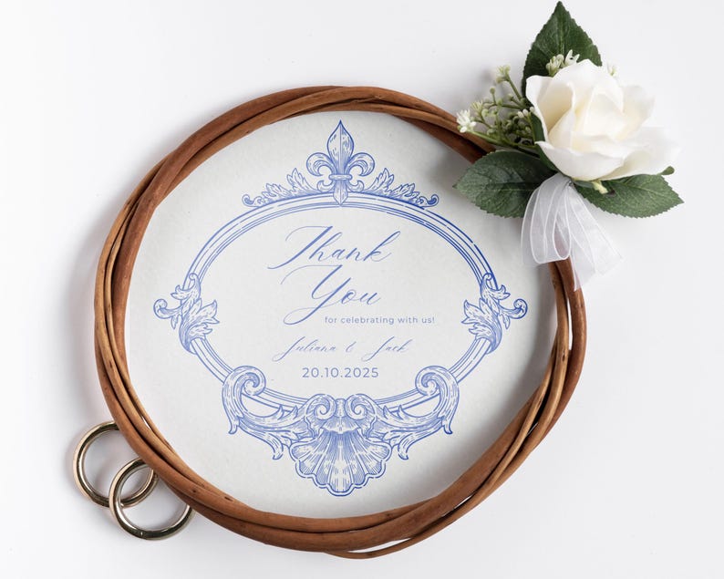 Puede incluir: Un recuerdo de boda con un dise&ntilde;o floral azul y la frase "Thank You". Una corona trenzada marr&oacute;n rodea el dise&ntilde;o, con una rosa blanca y vegetaci&oacute;n adjunta. Se ven dos anillos dorados.