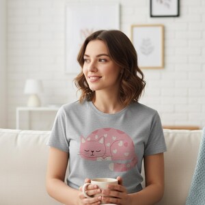 Pode incluir: Uma camiseta cinza com um desenho de gato rosa com detalhes de cora&ccedil;&otilde;es brancos. O gato est&aacute; enrolado e dormindo. A camiseta &eacute; de manga curta e feita de um material macio.