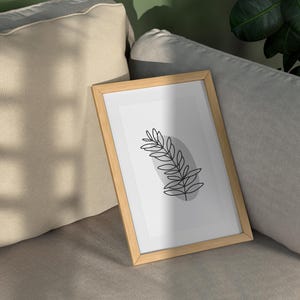 May include: A framed print of a simple line drawing of a leaf in black on a white background. The frame is light brown wood and is sitting on a light gray couch.