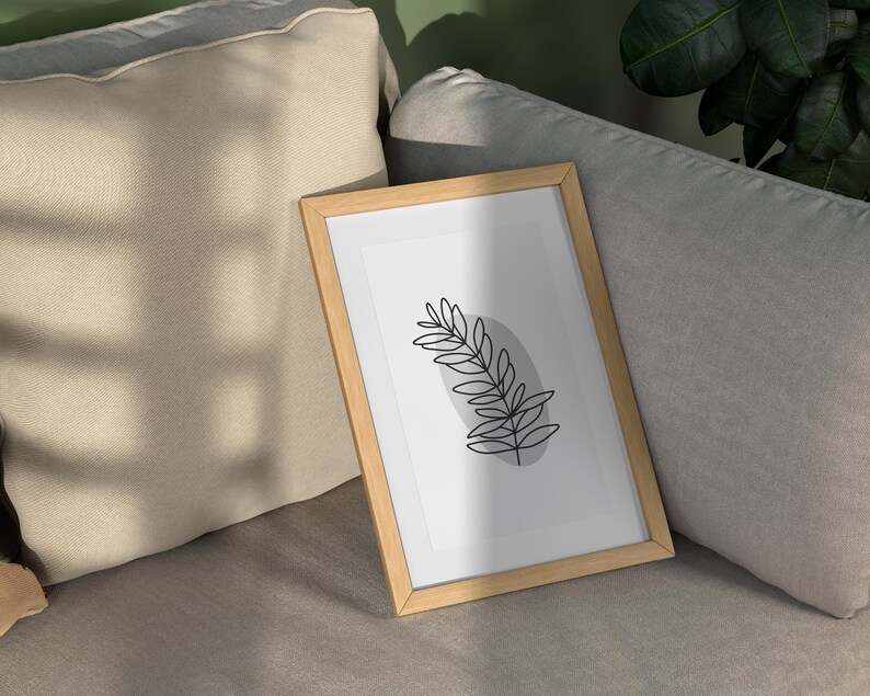 May include: A framed print of a simple line drawing of a leaf in black on a white background. The frame is light brown wood and is sitting on a light gray couch.