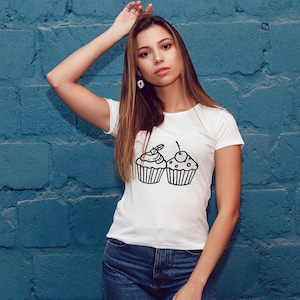 K&ouml;nnte beinhalten: Wei&szlig;es T-Shirt mit einer schwarzen Strichzeichnung von zwei Cupcakes. Einer hat eine Kirsche, der andere ein Wirbeldesign. Das Shirt wird mit blauen Jeans kombiniert. Der Hintergrund ist eine blaue Backsteinmauer.