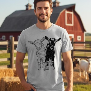 May include: A light grey t-shirt with a black and white illustration of two young goats. The goats are side by side. The background shows a red barn, a wooden fence, and a field with hay bales.