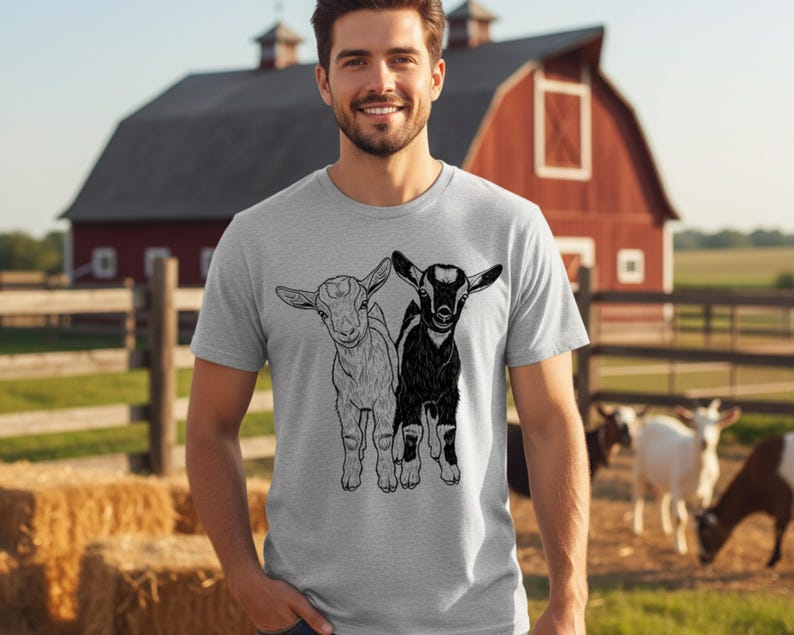 May include: A light grey t-shirt with a black and white illustration of two young goats. The goats are side by side. The background shows a red barn, a wooden fence, and a field with hay bales.