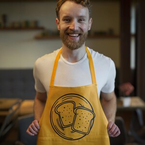 K&ouml;nnte beinhalten: Senfgelbe Sch&uuml;rze mit einer schwarzen Illustration von zwei Toastscheiben auf einem Teller. Die Sch&uuml;rze hat eine Tasche und gelbe Tr&auml;ger. Die Person auf dem Bild tr&auml;gt ein wei&szlig;es T-Shirt.