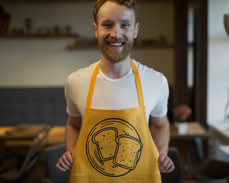 K&ouml;nnte beinhalten: Senfgelbe Sch&uuml;rze mit einer schwarzen Illustration von zwei Toastscheiben auf einem Teller. Die Sch&uuml;rze hat eine Tasche und gelbe Tr&auml;ger. Die Person auf dem Bild tr&auml;gt ein wei&szlig;es T-Shirt.