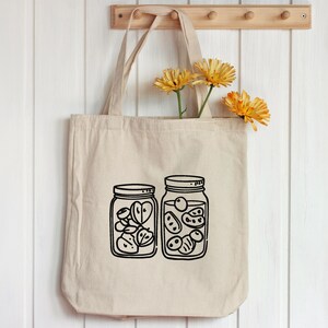 K&ouml;nnte beinhalten: Beige Canvas-Tragetasche mit schwarzer Strichzeichnung von zwei mit Fr&uuml;chten gef&uuml;llten Einmachgl&auml;sern. Die Tasche h&auml;ngt an einer Holzleiste mit gelben Blumen. Die Gl&auml;ser sind mit geschnittenem Obst gef&uuml;llt.