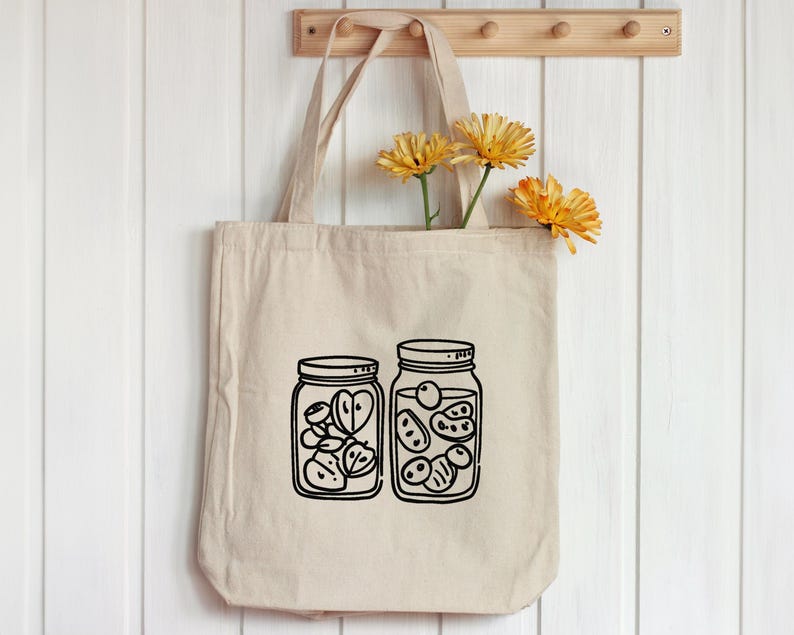 K&ouml;nnte beinhalten: Beige Canvas-Tragetasche mit schwarzer Strichzeichnung von zwei mit Fr&uuml;chten gef&uuml;llten Einmachgl&auml;sern. Die Tasche h&auml;ngt an einer Holzleiste mit gelben Blumen. Die Gl&auml;ser sind mit geschnittenem Obst gef&uuml;llt.