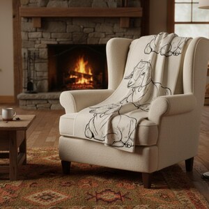 May include: A cream armchair with a throw blanket featuring a black goat illustration. The chair is positioned in front of a lit fireplace, creating a warm, inviting scene. The blanket is draped over the chair. The room has a rustic feel.
