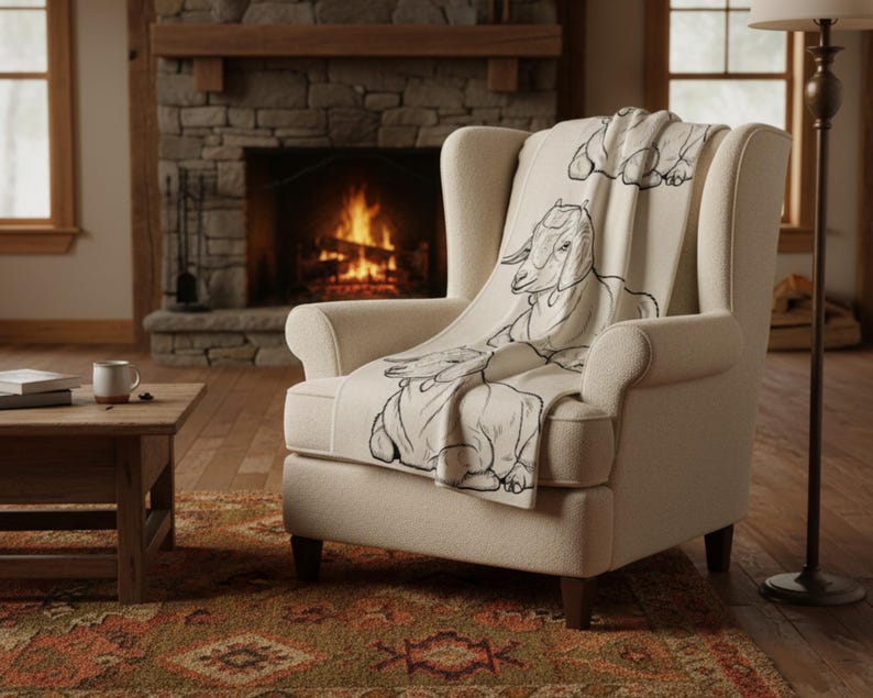 May include: A cream armchair with a throw blanket featuring a black goat illustration. The chair is positioned in front of a lit fireplace, creating a warm, inviting scene. The blanket is draped over the chair. The room has a rustic feel.