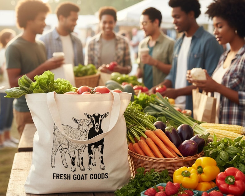 May include: A cream canvas tote bag filled with fresh produce at a market stall. The bag has a goat illustration and reads "FRESH GOAT GOODS". The stall displays tomatoes, lettuce, carrots, peppers, and strawberries. People are browsing the fresh produce.