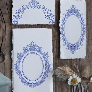 Puede incluir: Tres trozos de papel blanco con dise&ntilde;os de marcos azules ornamentados. Los dise&ntilde;os incluyen marcos ovalados y elementos florales decorativos. El papel tiene un borde irregular. Un peine con flores y plantas secas est&aacute; en la esquina inferior derecha.