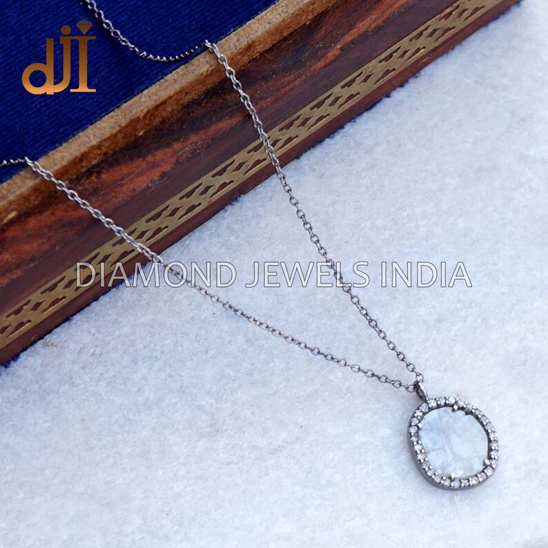 May include: A silver chain necklace with a round pendant featuring a white stone surrounded by smaller diamonds. The pendant is on a white surface with a wooden box in the background. The text "DIAMOND JEWELS INDIA" is visible below the box.