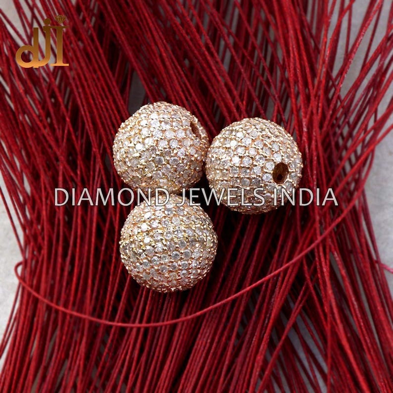 K&ouml;nnte beinhalten: Drei runde, goldfarbene Perlen, besetzt mit kleinen, funkelnden Steinen. Sie liegen auf einem Hintergrund aus roten F&auml;den. Der wei&szlig;e Text lautet "DIAMOND JEWELS INDIA".
