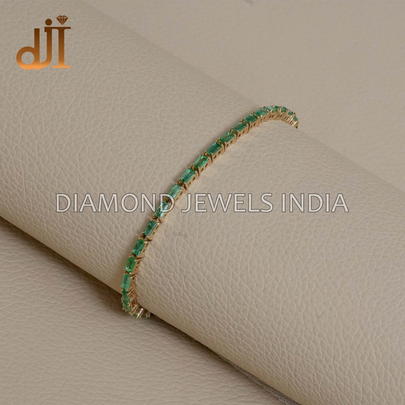 May include: A gold bracelet with a row of emerald green rectangular gemstones. The bracelet is on a light brown surface. DIAMOND JEWELS INDIA is printed on the surface.