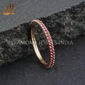 Puede incluir: Un anillo de banda de oro con una fila de pequeñas piedras preciosas rojas. El anillo está sobre una superficie texturizada gris oscuro.