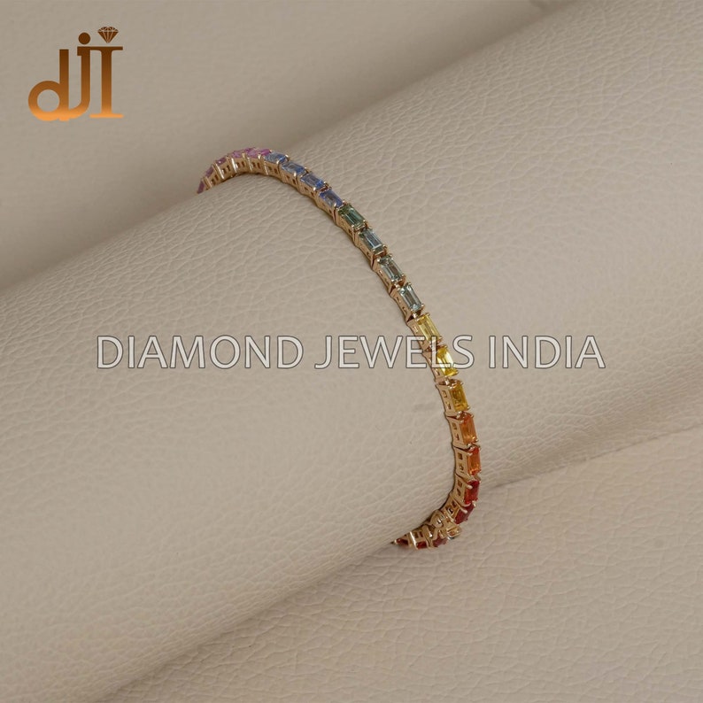 May include: A gold bracelet with a rainbow of baguette-cut gemstones. The bracelet is on a light brown surface. DIAMOND JEWELS INDIA is printed on the surface.