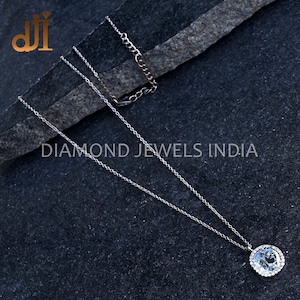 May include: A silver chain necklace with a round pendant featuring a large clear gemstone surrounded by smaller clear gemstones. The pendant is set in a silver bezel setting. The necklace is on a dark gray textured surface. DIAMOND JEWELS INDIA is printed on the surface.