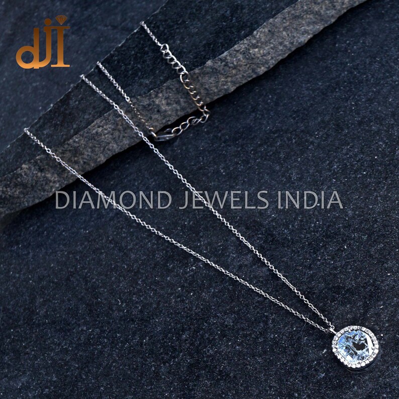 May include: A silver chain necklace with a round pendant featuring a large clear gemstone surrounded by smaller clear gemstones. The pendant is set in a silver bezel setting. The necklace is on a dark gray textured surface. DIAMOND JEWELS INDIA is printed on the surface.