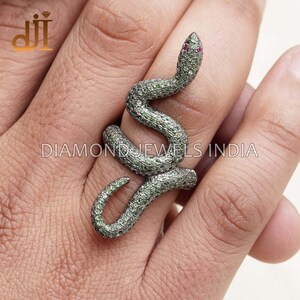 May include: A silver snake ring with green gemstones and a red gemstone for the eyes. The ring is on a person's finger. DIAMOND JEWELS INDIA is printed on the ring.