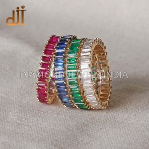 Può includere: Quattro anelli d'oro con pietre preziose di diversi colori. Gli anelli sono impilati uno sopra l'altro. Le pietre preziose sono rosse, blu, verdi e trasparenti.  DIAMOND JEWELS INDIA è stampato sulla superficie sotto gli anelli.