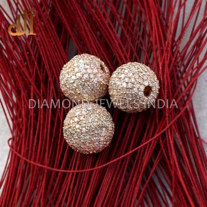 K&ouml;nnte beinhalten: Drei goldfarbene, kugelf&ouml;rmige Perlen, die mit kleinen, klaren Steinen besetzt sind. Die Perlen sind auf einem Hintergrund aus roten F&auml;den angeordnet. Der Text "DIAMOND JEWELS INDIA" ist in Wei&szlig; zu sehen.