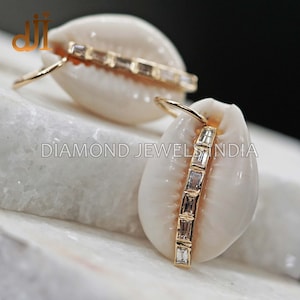 May include: Gold earrings featuring a white seashell design with a row of clear baguette-shaped stones.