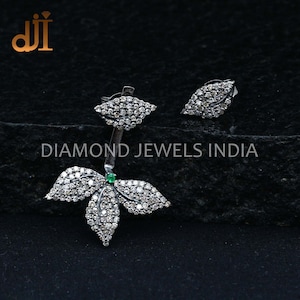 May include: A silver-toned leaf-shaped earring set with a matching stud. The earrings are encrusted with small, sparkling stones. A small green stone is set in the center of the larger earring. The text "DIAMOND JEWELS INDIA" is visible.