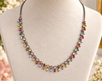 May include: A dark metal chain necklace featuring a series of alternating round and baguette-cut multicolored gemstones. The stones are set in a repeating pattern of red, yellow, green, blue, and purple hues, dangling from a thin, dark link chain.