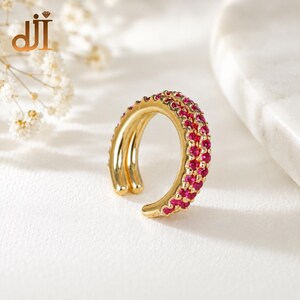May include: A gold ear cuff featuring a double-band design with one band encrusted with small, vibrant pink stones. The piece has an open, circular shape, showcasing a polished metallic finish and a row of sparkling, round-cut pink gemstones.