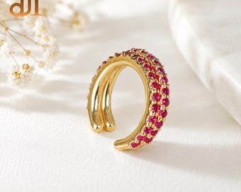 May include: A gold ear cuff featuring a double-band design with one band encrusted with small, vibrant pink stones. The piece has an open, circular shape, showcasing a polished metallic finish and a row of sparkling, round-cut pink gemstones.