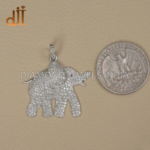 Puede incluir: Un colgante de elefante plateado incrustado con pequeñas piedras preciosas transparentes. El elefante tiene un ojo de piedra preciosa roja y está al lado de una moneda. De Diamond Jewels India.