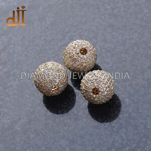 K&ouml;nnte beinhalten: Drei runde, goldfarbene Perlen, besetzt mit kleinen, klaren Edelsteinen. Die Perlen liegen auf einer dunkelgrauen Oberfl&auml;che, mit dem Text "DIAMOND JEWELS INDIA" im Hintergrund.