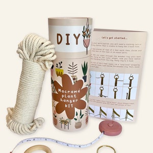 May include: A DIY macrame plant hanger kit with a beige rope, a wooden ring, a pink measuring tape, a gold hook, and instructions on a beige card with illustrations of plants in pots. The card reads "Let's get started..." and "Macrame Plant Hanger Kit".
