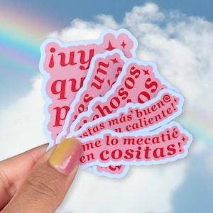 May include: A hand holds a stack of pink stickers with red text. The stickers feature phrases like "juy," "un," and "stas más buen@ n caliente!" Each sticker has a light blue border. The background shows a sky with clouds and a rainbow.