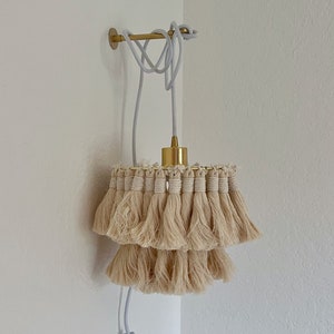 Könnte beinhalten: Eine beige Quasten-Pendelleuchte mit einem goldenen Metallkabel und einem weißen Kabel-Schalter. Die Leuchte ist an einer weißen Wand montiert.
