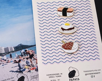 May include: Set of three gold enamel pins featuring Hawaiian food: a Spam Musubi, a Loco Moco, and a plate of Kalua Pig with rice. The pins are on a white card with a blue and pink wave pattern. The card also includes the text "Hawaii Foodie Pins", "Homegrown in Small Town AIEA, HI", and "Papaya Party".