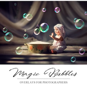 May include: A baby in a white dress and white hat reaches for iridescent soap bubbles floating in the air. The bubbles are various sizes and are against a backdrop of a white curtain and a wooden floor.