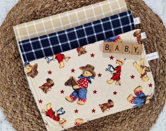 Op de afbeelding: Drie spuugdoeken met een cowboythema. De bovenste doek is beige met witte vierkanten, de middelste doek is donkerblauw met witte vierkanten en de onderste doek is wit met een rood sterpatroon en bruine teddyberen die op paarden rijden. De tekst "BABY" is gespeld met lettertegels.