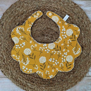 May include: Mustard yellow baby bib with a floral pattern of white flowers and brown stems. The bib has a scalloped edge and fastens with two wooden buttons. The bib is made of soft fabric.