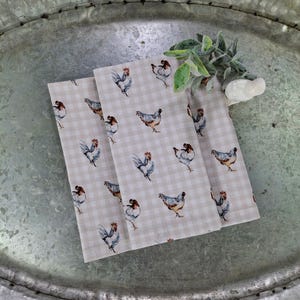 May include: Two beige tea towels with a gingham pattern and a watercolor design of roosters and hens.