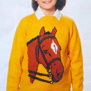 May include: A yellow knitted sweater with a brown horse head design. The sweater has a white collar and is worn by a young person wearing a white beret.
