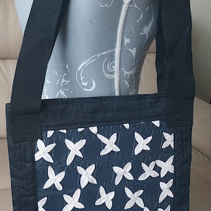 May include: A black crossbody bag with a wide shoulder strap. The bag features a navy blue front panel with a repeating white floral pattern. The edges and strap are black.