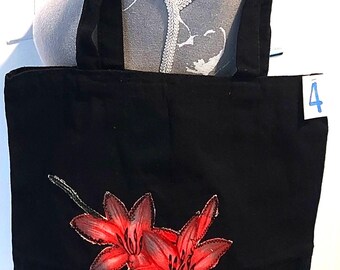 May include: Black tote bag with a vibrant red floral design. The bag features a pair of stylized red lilies with green stems. The tote has black handles and is displayed on a mannequin.