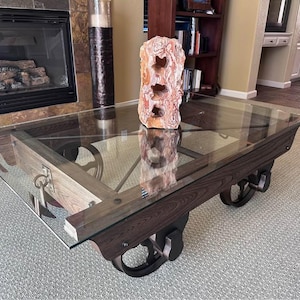 May include: A rectangular coffee table featuring a clear glass top supported by a dark wood frame with metal accents. The base includes four black metal wheels and a metal ring handle on the side. A tall, textured, orange and white stone sculpture sits on top.