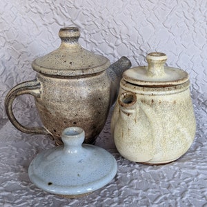 May include: Three ceramic teapots with lids. One is a light brown with a handle and spout, the second is a cream color, and the third is a light blue lid. The teapots are on a textured white surface.