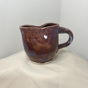 Puede incluir: Una taza de cerámica hecha a mano con un borde en forma de corazón, que presenta un esmalte marrón brillante con sutiles tonos azules y un asa gruesa y curva. La taza tiene una forma orgánica ligeramente irregular y un acabado superficial liso.