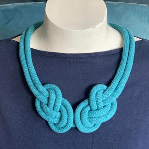 May include: A turquoise blue knotted necklace displayed on a white mannequin head. The necklace is made of a thick, woven material and features two large knots at the front. The background is a dark blue top.