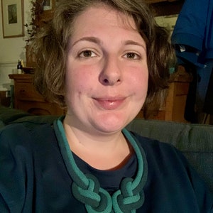 May include: A close-up shot of a teal and dark green knotted necklace. The necklace is made of a thick, rope-like material and features two large, symmetrical knots at the center. The person wearing the necklace is wearing a dark teal shirt.