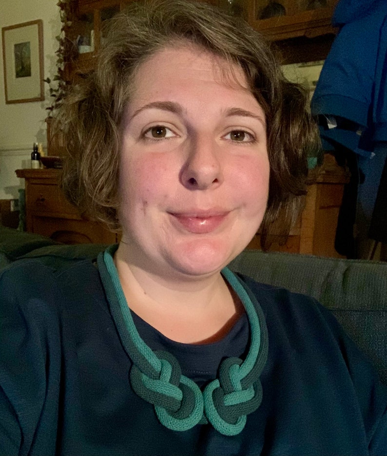 May include: A close-up shot of a teal and dark green knotted necklace. The necklace is made of a thick, rope-like material and features two large, symmetrical knots at the center. The person wearing the necklace is wearing a dark teal shirt.
