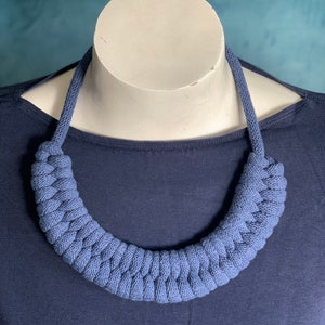 Op de afbeelding: Een blauwe gebreide ketting die wordt getoond op een witte mannequinhoofd. De ketting heeft een dik, gevlochten ontwerp in een effen blauwe kleur. De mannequin staat tegen een donkerblauwe top.